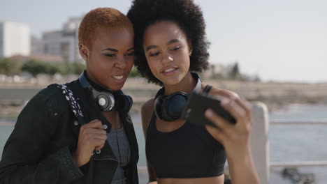 portrait of two beautiful african american woman uing phone on beachfront posing taking selfie