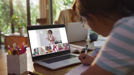 Schoolgirl-using-laptop-for-online-lesson-at-home,-with-diverse-teacher-and-class-on-screen