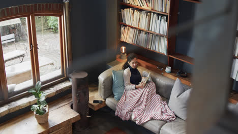 biracial woman lying on sofa under blanket and reading book at home, slow motion