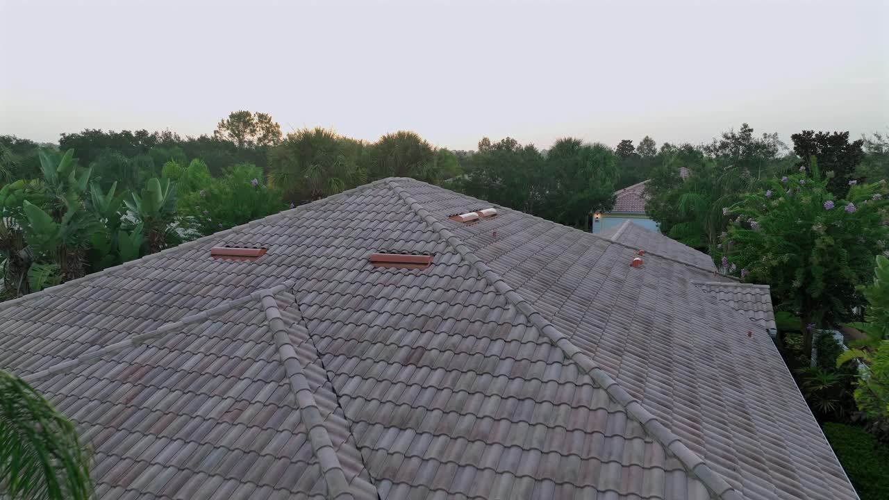 Suburban Florida home with tiled roof and screened lanai, surrounded by tropical landscaping and palm trees. Single-story architecture typical of hurricane-prone coastal areas. Aerial approaching