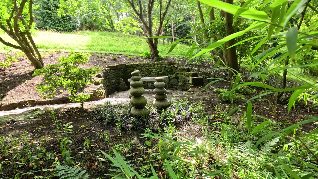 Camera slowly pans through lush greenery, revealing a stacked stone sculpture in a tranquil botanical garden under bright, natural daylight