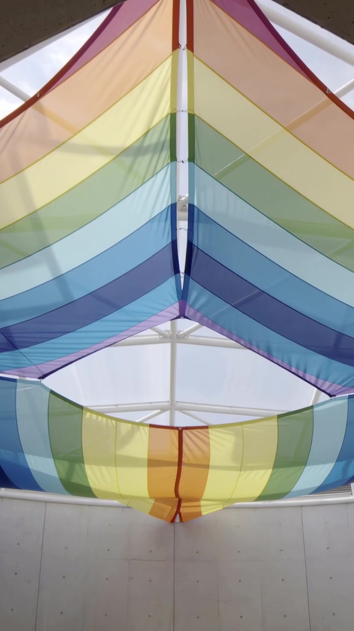 Video verticale: Luce solare attraverso il lucernario che illumina una tettoia arcobaleno nell'atrio, con i pannelli che ondeggiano dolcemente.
