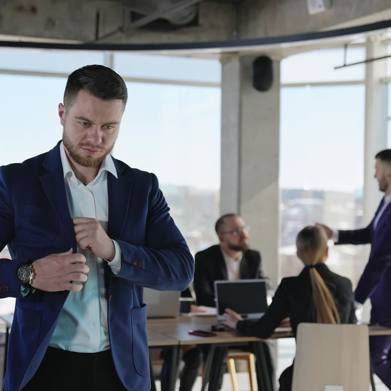 Confident young entrepreneur looks at his phone and then puts it into the pocket. Handsome man in a suit in the office. Team of colleagues at the background