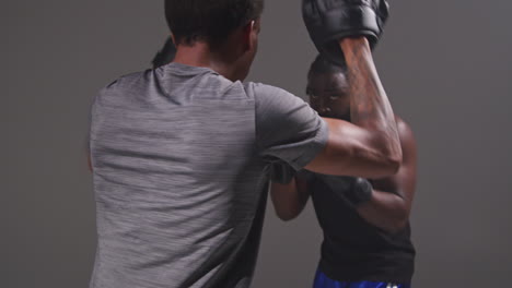 Studio-Shot-Of-Male-Boxer-Sparring-Working-Out-With-Trainer-Wearing-Punch-Mitts-Or-Gloves-Practising-For-Fight