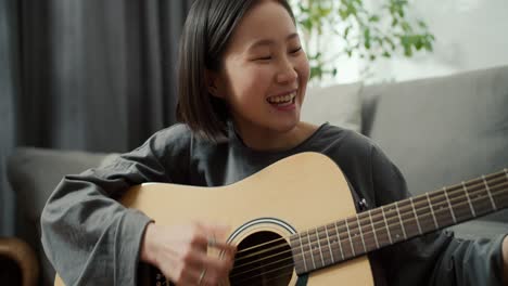 Chica-Asiática-Tocando-Instrumento-Musical-De-Guitarra-Acústica-En-Casa,-Sentada-En-El-Suelo,-De-Cerca