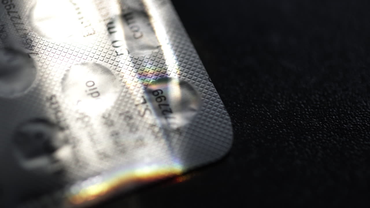 Close up of multicolored light rays dancing across pill blister pack surface, casting iridescent reflections against contrasting dark background, highlighting pharmaceutical product details