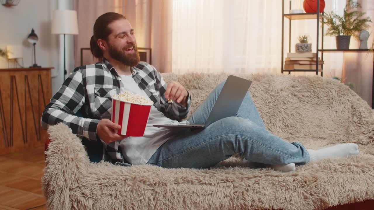 Young man relaxing at home sofa eating popcorn bowl laptop laughter video comedic fun enjoyment