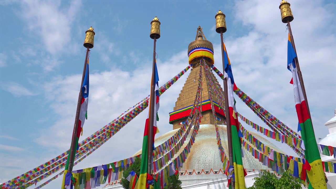 Ripresa in movimento dal basso della maestosa Stupa di Boudhanath circondata da alti pali di bandiere di preghiera in ottone e colorati stendardi sventolanti sotto un cielo parzialmente nuvoloso a Kathmandu, Nepal