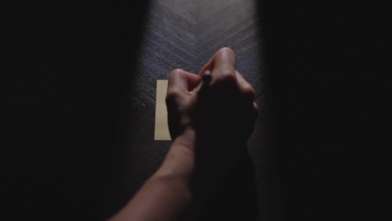 Slow motion close up footage of a hand writing out "Happy 4th of July" on a paper note and placing it on a table. Shot in dramatic lighting for an impressive title or transition