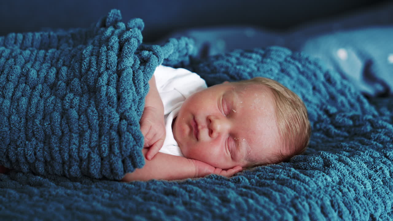 Adorable Caucasian newborn peacefully sleeping. Cute baby put a hand under his cheek sleeps on his side covered in blue woven blanket. Close up.