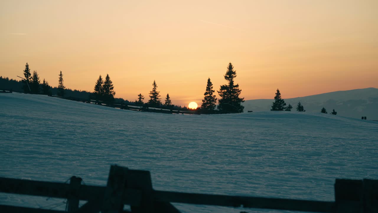Tramonti dorati su un campo montano innevato con staccionata in legno e alberi.