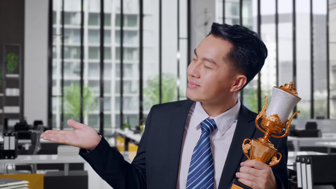 Close Up Of Asian Business Man In A Suit And Tie With A Gold Medal Looking At A Gold Trophy In His Hands, Smiling, And Pointing To Side In Office