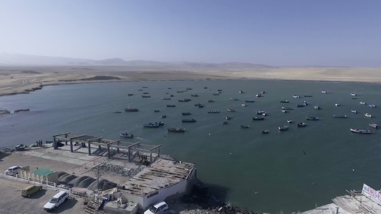 Long aerial drone shot of the Peruvian coastline of a beach with many small fishing boats with birds and seagulls flying around