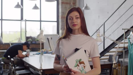 portrait of beautiful young fashion designer holding her sketchbook and smiling at camera in sewing workshop while her colleagues working in the background