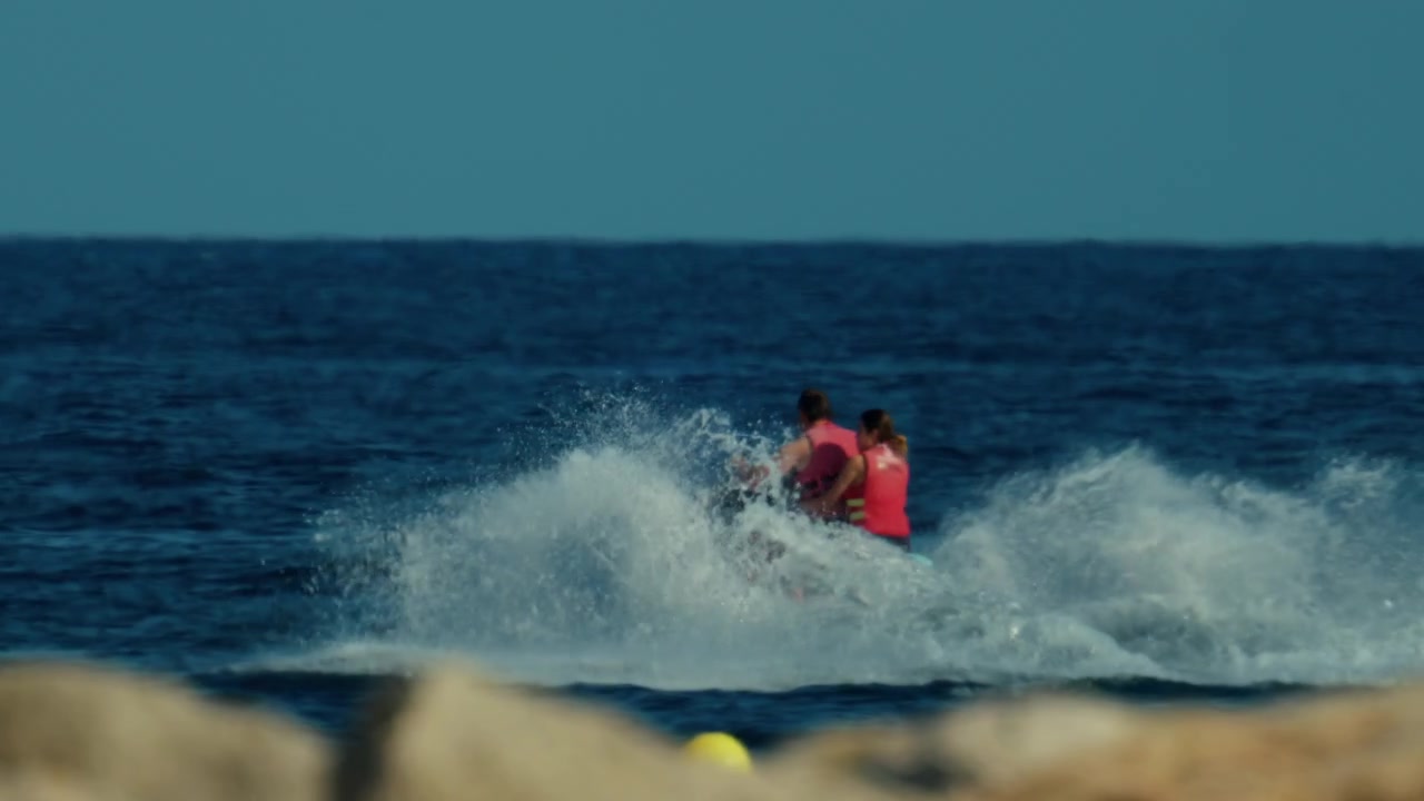 To personer suser afsted på en jetski over det blå hav og skaber hvide sprøjt. En gul bøje flyder i forgrunden, Antibes, Frankrig
