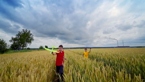 Children playing in farmland. Boys running with toy planes in the wheat field in summer. Brothers having fun outdoors under cloudy sky. Front view.
