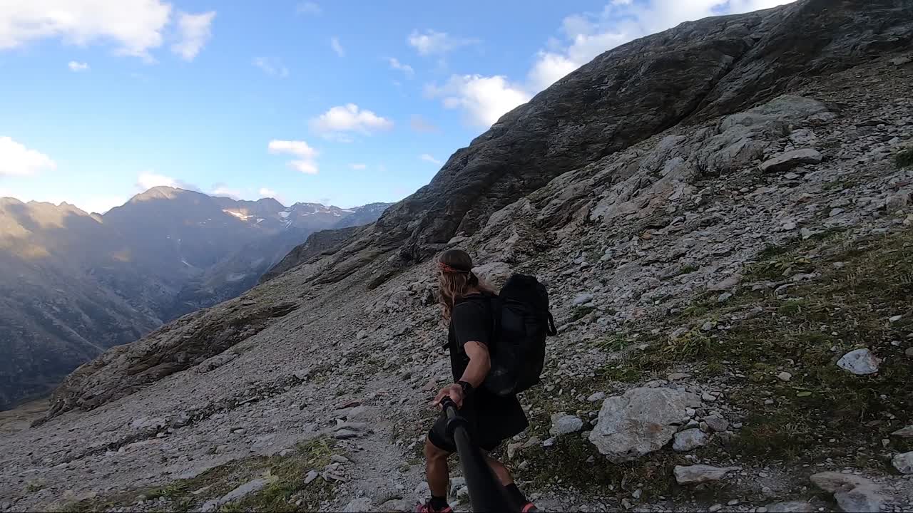 un hombre joven y en forma con el pelo largo y una mochila grande está trotando y corriendo con cuidado por un camino en las montañas llenas de rocas y piedras