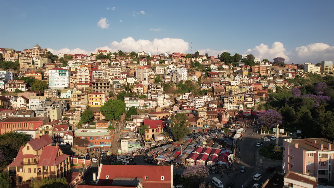 A short aerial clip revealing the edge of Ambohijatovo Market in Antananarivo, Madagascar, with jacaranda trees blooming in the lower frame and layers of hillside homes above