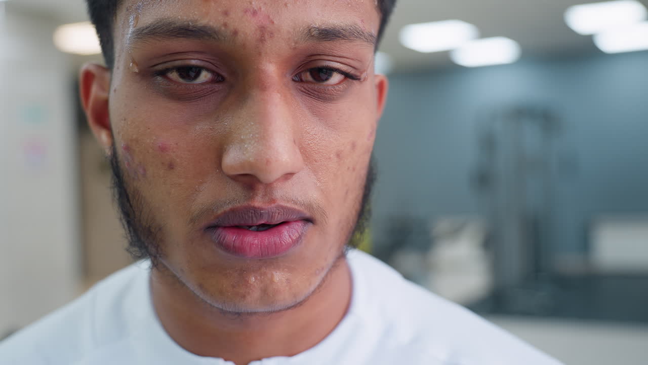 close up of exhausted man taking break after intense treadmill run, sweat beads on forehead and cheeks, head tilted down in recovery, blurred gym equipment and warm bokeh lighting in background