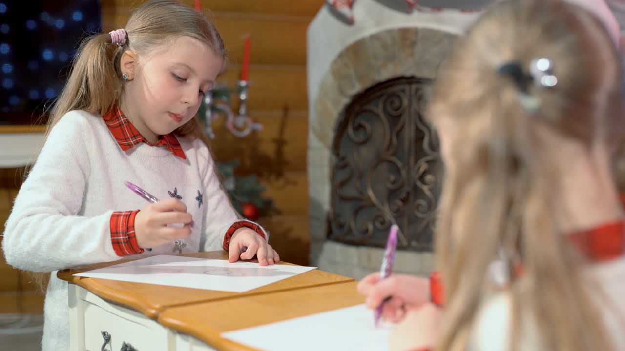 Little girls draw at the table near the fireplace and the Christmas tree. Two sisters write a letter to Santa Claus before the new year