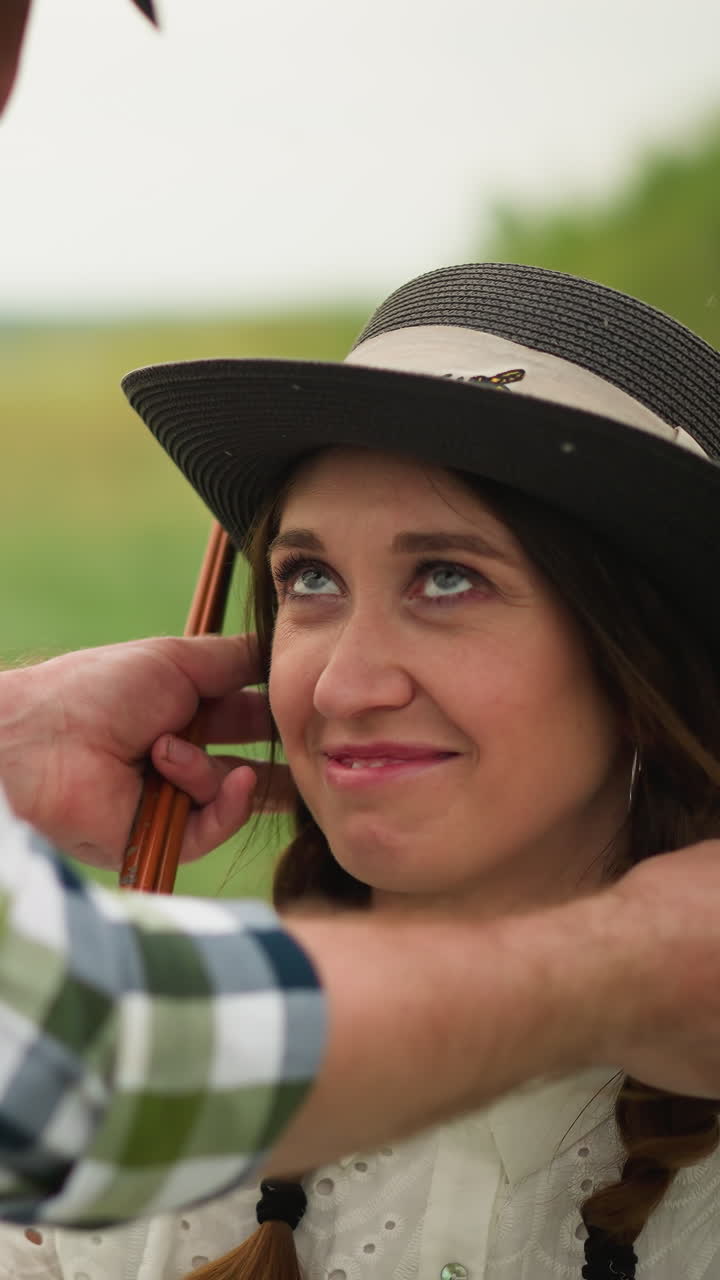 artista en una camisa a cuadros ajusta suavemente el sombrero de una mujer sonriente con el cabello trenzado, con un vestido blanco, sentado en un campo verde