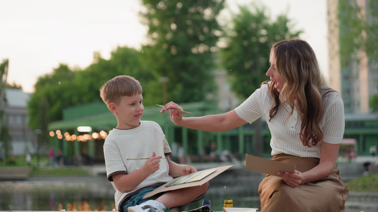 caregiver and son seated on wooden dock by pool at sunset, mother playfully touches son s cheek with paintbrush while he smiles and paints on board under warm evening glow with water reflections