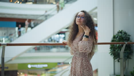 mujer en vestido vintage se encuentra cerca de la barandilla de hierro, ajustando su cabello pensativamente en un centro comercial brillante y espacioso, el borrón de fondo muestra a la gente moviéndose en escaleras mecánicas