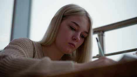 mujer joven enfocada escribiendo con una expresión seria, profundamente en pensamiento, sosteniendo una pluma sobre un cuaderno, con fondo borroso con barandilla de metal y iluminación suave