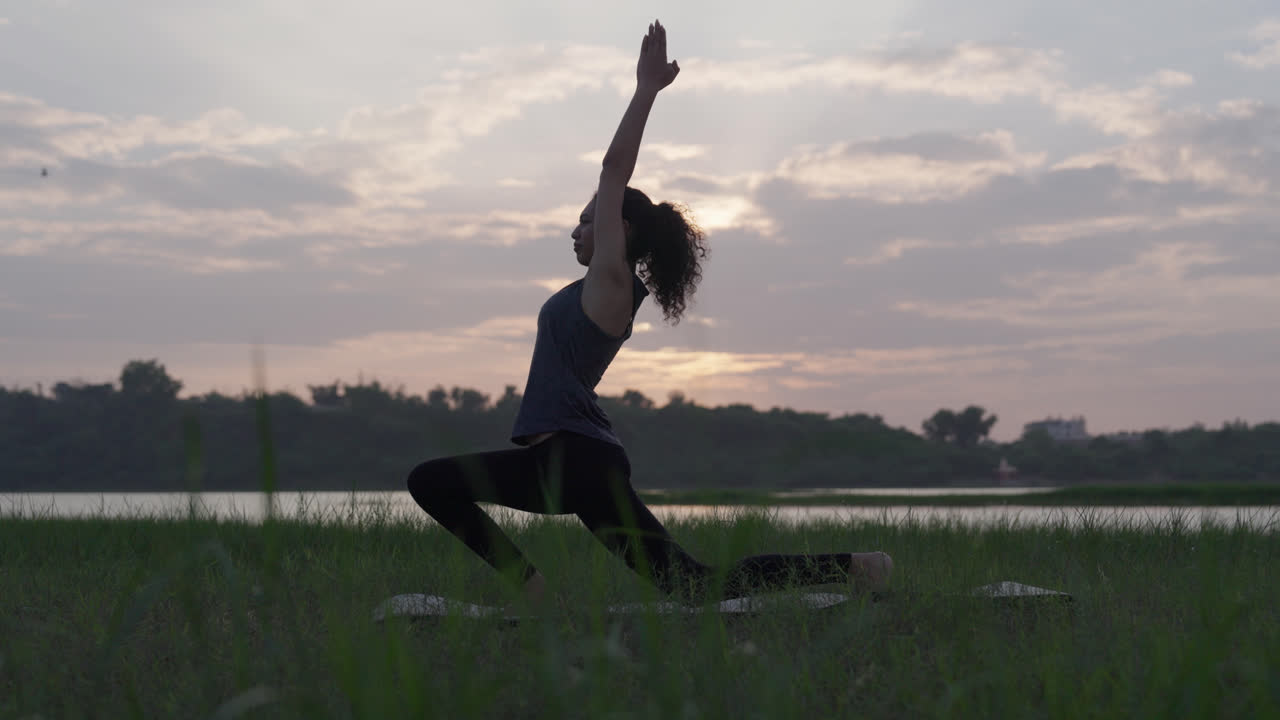 Un rallentatore di una donna dell'Asia meridionale in posizione yoga di affondo basso (Anjaneyasana) con le braccia alzate, al tramonto vicino a un lago