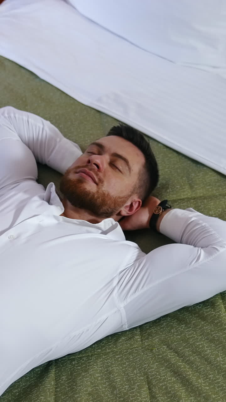Young businessman relaxing on bed. Handsome man in white shirt is lying on bed enjoying rest alone in a hotel room. Vacation during business trip. Vertical video