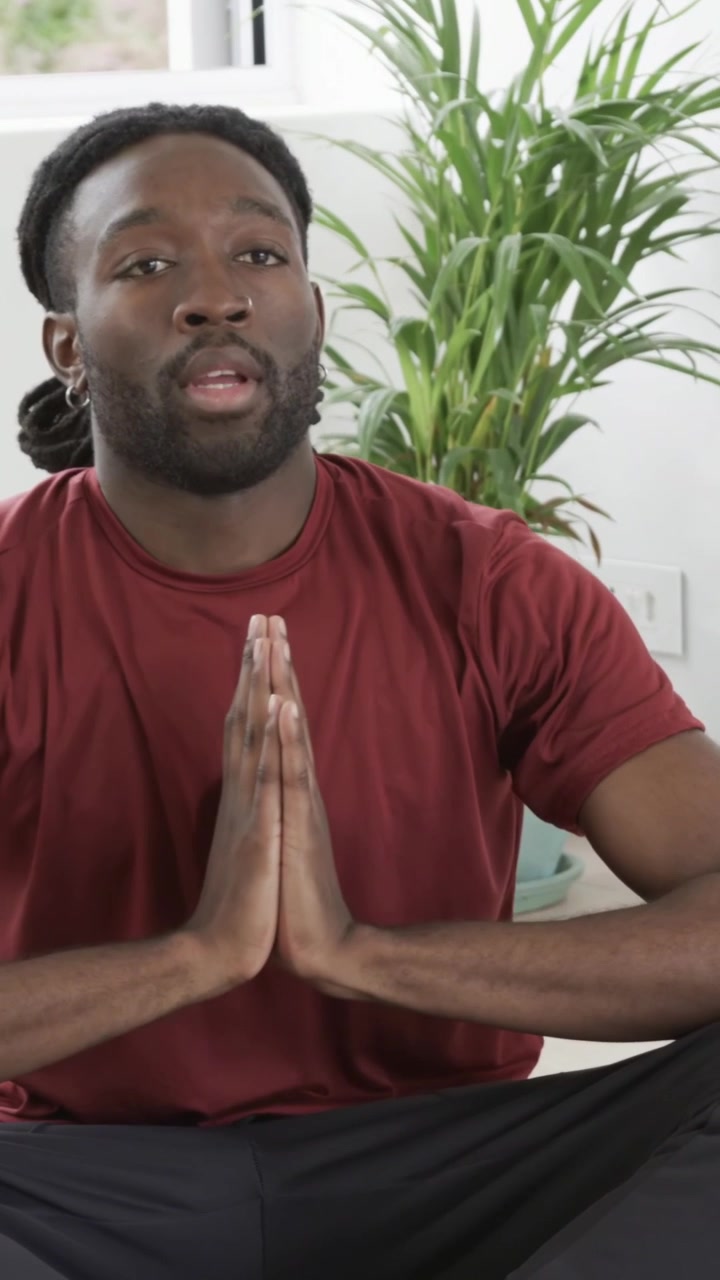 Vertical video: Raising adult male wearing maroon tee doing yoga on mat at home by window, plant