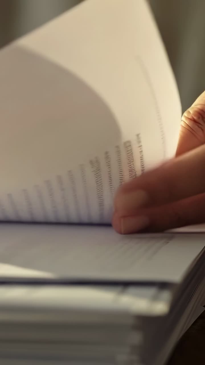 Vídeo vertical: Mãos se movendo revisando e virando páginas impressas na mesa, com uma cortina ao fundo.