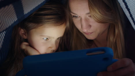 happy mother and daughter using tablet computer under blanket playing games on touchscreen technology relaxing having fun before bedtime