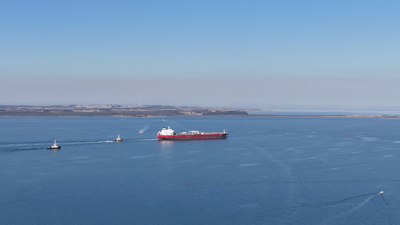 A large cargo ship navigates through serene waters, accompanied by two tug boats for assistance. The setting showcases bright blue skies and tranquility as the vessels move seamlessly.