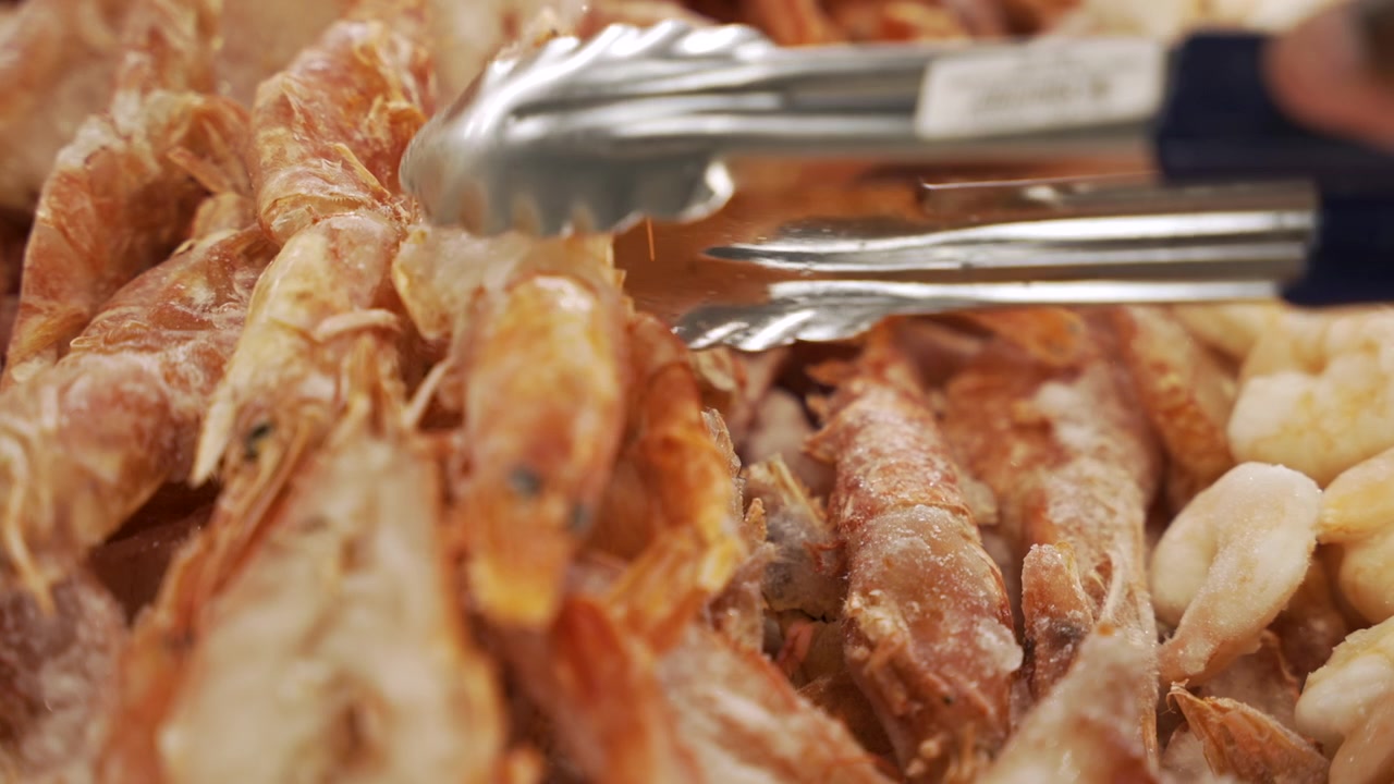 Serving tongs collect frozen shell-on prawns at a self-serve grocery bin beside peeled shrimp pieces in a bright retail food store.