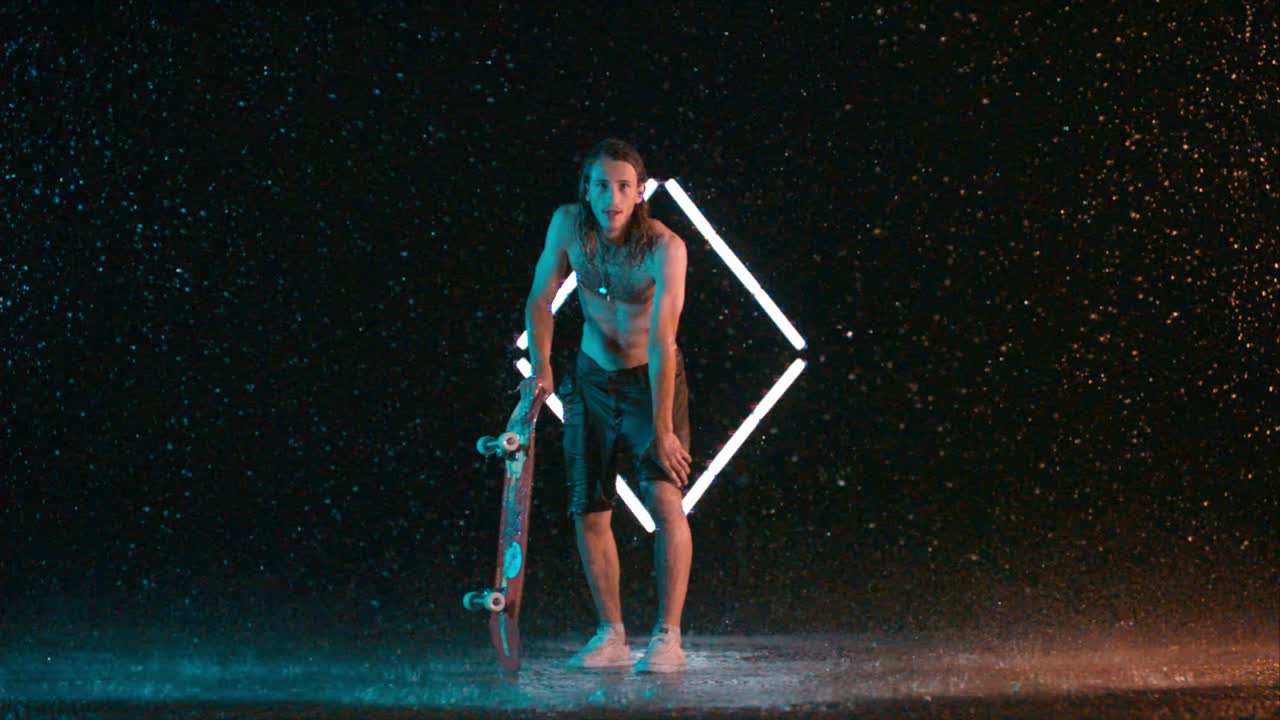 Caucasian male skateboarder holding board in rain and looking at camera, slow motion