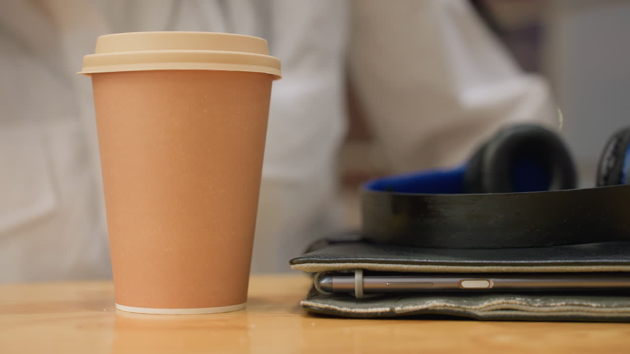 Close up of woman manicured hand gently placing coffee cup on wooden table beside blurred tablet and headphones, with glowing city bokeh lights through glass panel in soft focus