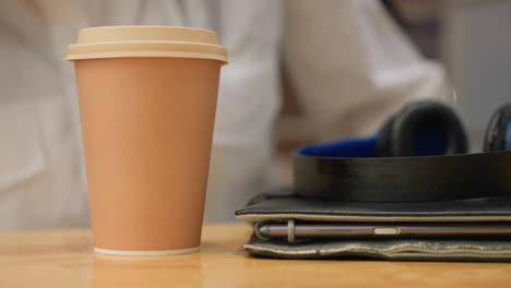 close up of woman manicured hand gently placing coffee cup on wooden table beside blurred tablet and headphones, with glowing city bokeh lights through glass panel in soft focus