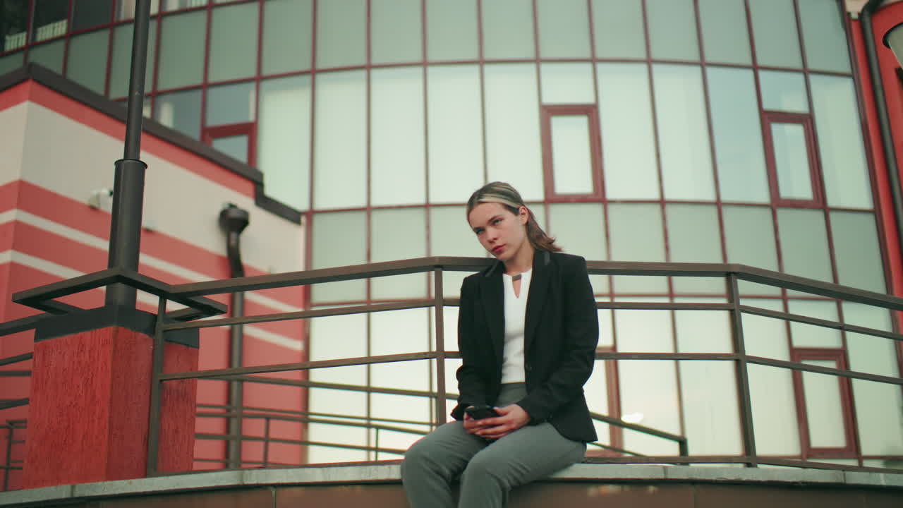 Reflective woman seated in solitude on fence with legs crossed at ankle, staring thoughtfully at phone in hand, dressed in professional outfit, surrounded by modern building and lampposts in urban setting