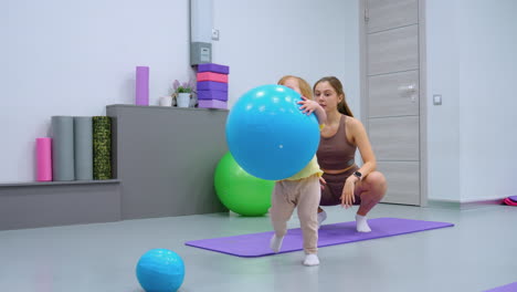 playful toddler grabs blue balloon from squatting mother, who shakes her head in gentle disagreement, colorful gym background enhances lively scene