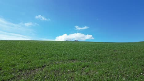 Flying-with-a-drone-over-a-green-meadow-with-some-spectacular-mountains-in-the-background-with-a-blue-sky-with-white-clouds