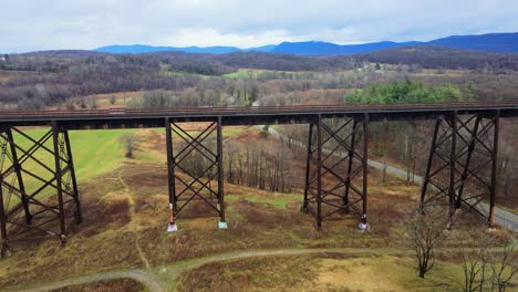 Aerial-drone-video-footage-of-a-train-bridge-viaduct-running-over-a-valley-in-the-Appalachain-Mountains-during-early-spring-on-a-cloud-day,-surrounded-by-mountains-and-farmland