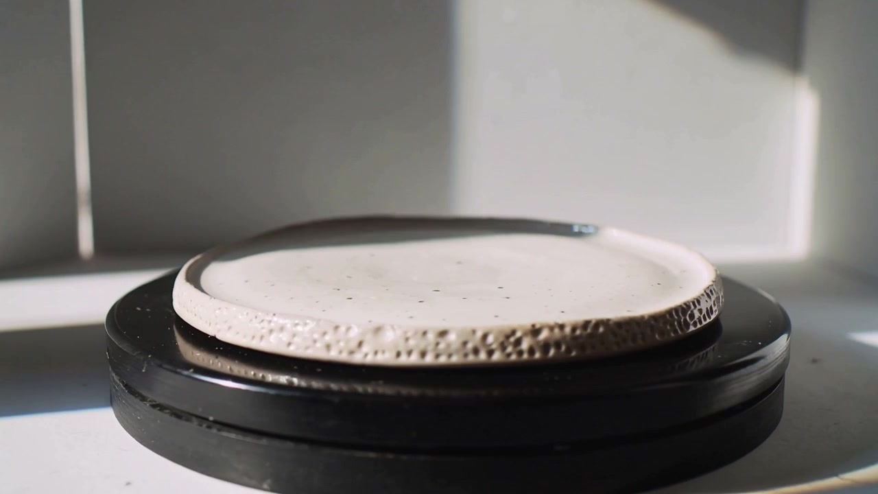 Işık yüzeyde gezinirken, bir bölmede çukurlu kenarını gösteren, siyah bir stand üzerindeki benekli seramik tabak dönüyor.