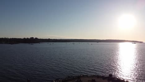 Luftpanorama-über-Sonnenuntergangsstrand-Bei-Heißer-Sonnenuntergangsreflexion-An-Einem-Frühen-Abend-Mit-Einer-Überführung-über-Den-Strandbereich,-Während-Frachtschiffe-Am-Hafen-Aufgereiht-Sind-Und-Segelboote-Sich-Darauf-Vorbereiten,-Für-Die-Nacht-Zu-Parken