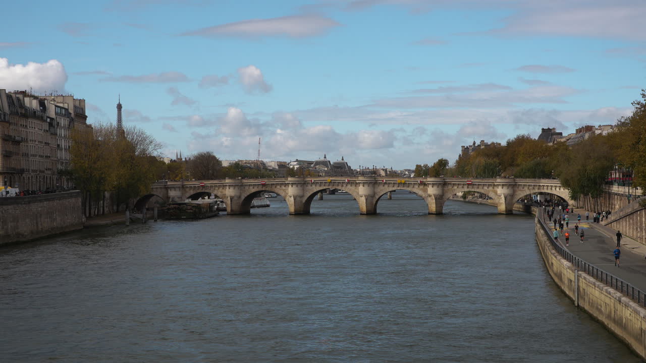 Clear daytime view of the Pont Neuf bridge spanning the Seine River in Paris with pedestrians walking along the quay and the Eiffel Tower visible in the distance under blue skies and gentle clouds