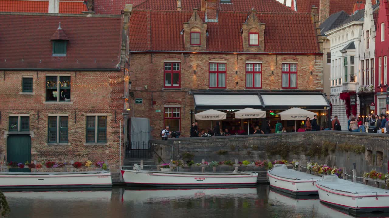 Crowds gather by medieval canal, boats docked, restaurants lit, soft evening light, steady camera