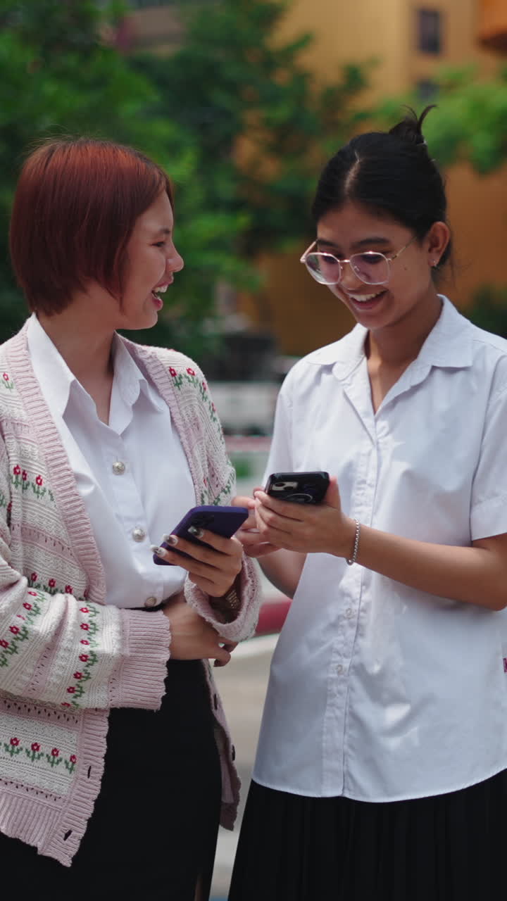 Two female students laughing and using cell phones on the street