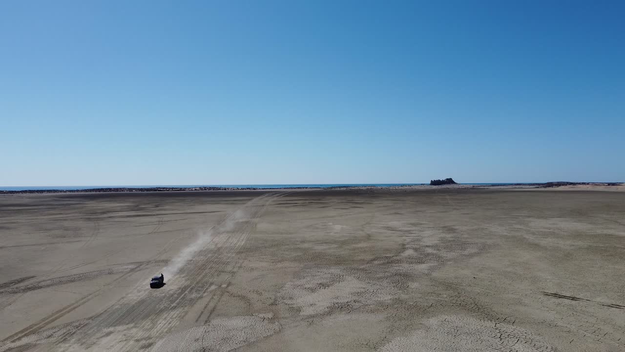 항공 드론 은  ⁇ 빛 이 많은 날 에 푸지 코, 발로치스탄 의 모래 사파트 해변 을 가로질러 운전 하는 관광 차량 의 뒤로 움직이는 장면 이다