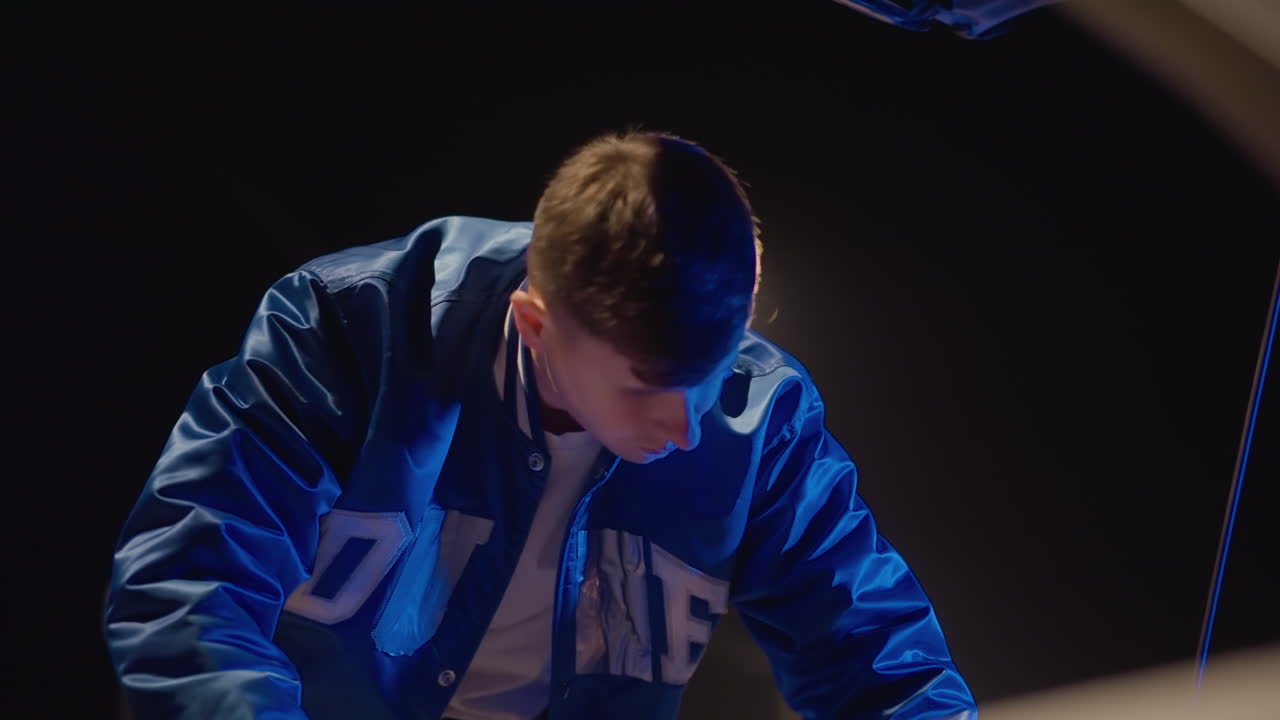 Conductor nocturno inspeccionando el capó del coche bajo la farola azul, joven asomándose al compartimento del motor, chaqueta universitaria captando reflejos fríos, ángulos cinematográficos en primer plano, expresión concentrada, urbano