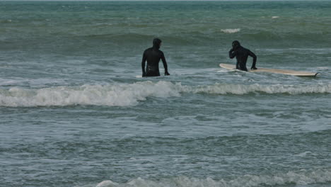 two surfers with insulated overalls enter the sea water with boards on a cold spring day.